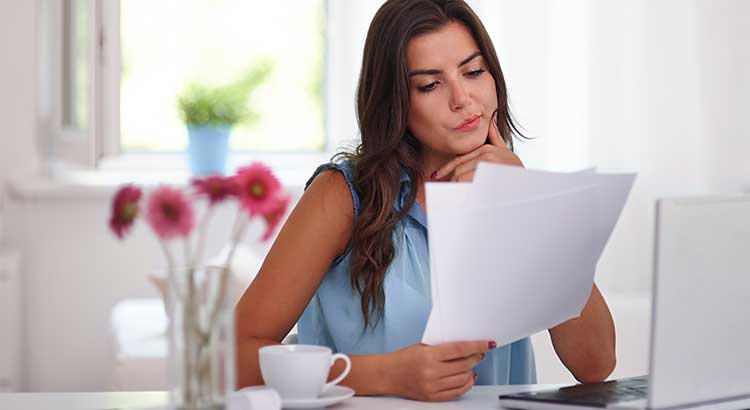 woman looks at papers and twists her mouth in ponderment, a cup of coffee and vase of flowers sit on the table.