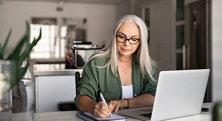 woman with long gray hair sits at desk writing with laptop open