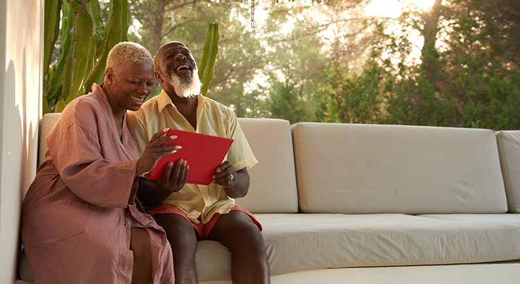older couple laughing looking at a tablet, sitting on a couch trees behind them.