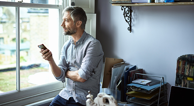 bearded man leans on home office desk with mobile phone in hand