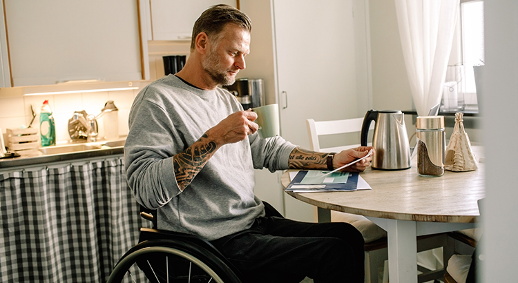 man in wheelchair at kitchen table reading and coffee cup in hand