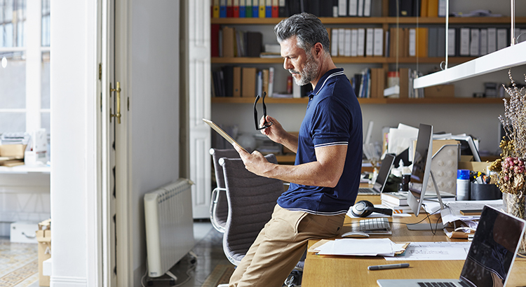 man holding reading glasses in hand looking at paper in other hand leaning on home office desk